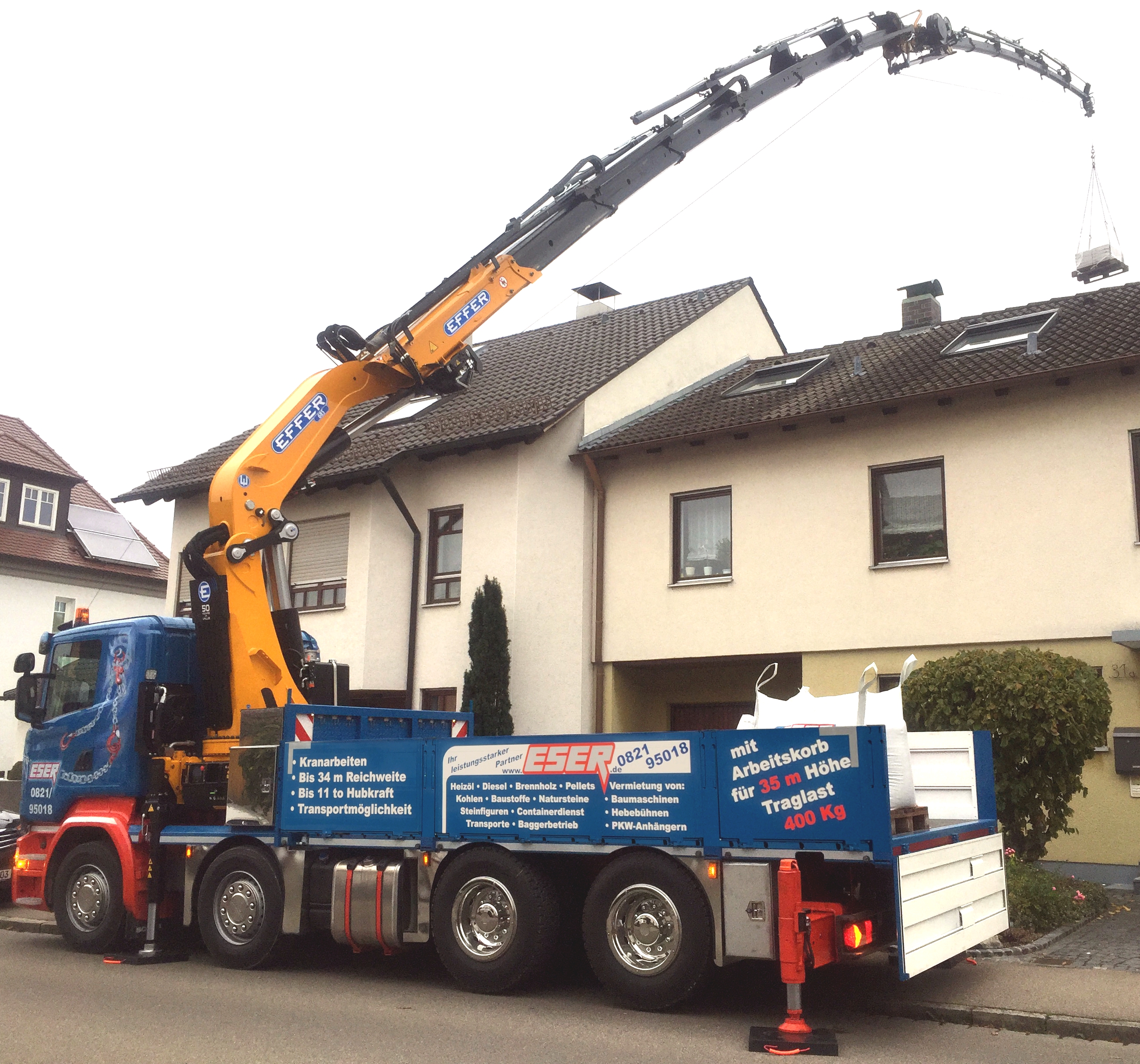 Das Foto zeigt Kranarbeit, bei der ein LKW mit Kranarm eine Last präzise über das Dach eines Hauses hebt. Eser in Augsburg bietet Spezialkrantransporte an und stellt verschiedene Kranservices zum Mieten bereit.
