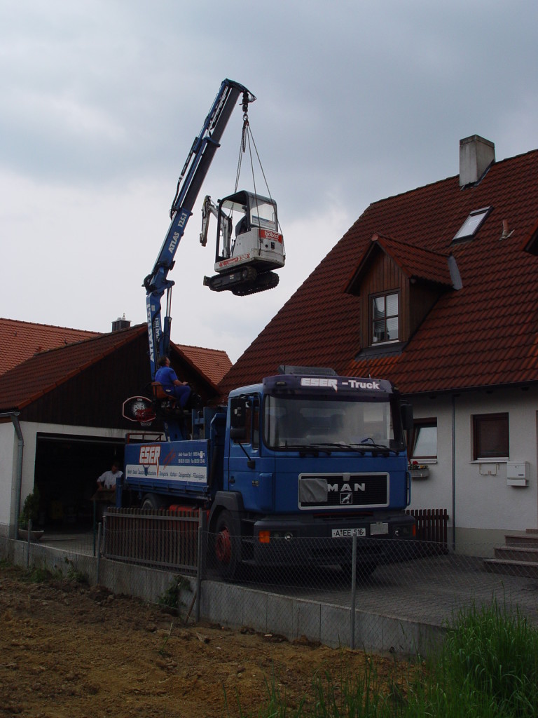 Das Foto zeigt Kranarbeit, bei der ein LKW mit Kranarm eine Baumaschine präzise über das Dach eines Hauses hebt. Eser in Augsburg bietet Spezialkrantransporte an und stellt verschiedene Kranservices zum Mieten bereit.
