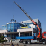 Das Foto zeigt Kranarbeit, bei der ein Lkw mit Kranarm eine Last auf das Dach eines Gebäudes hebt. Eser in Augsburg bietet Spezialkrantransporte an und stellt verschiedene Kranservices zum Mieten bereit