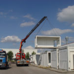 Das Foto zeigt Kranarbeit, bei der ein Lkw mit Kranarm einen Container präzise anhebt und an seinen Bestimmungsort bewegt. Eser in Augsburg bietet Spezialkrantransporte an und stellt verschiedene Kranservices zum Mieten bereit.