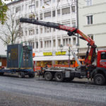 Das Foto zeigt Kranarbeit, bei der ein Lkw mit Kranarm eine Containerstruktur in der Innenstadt anhebt. Eser in Augsburg bietet Spezialkrantransporte an und stellt verschiedene Kranservices zum Mieten bereit.